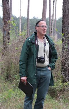 Don Bragg in Forest Service uniform in Kisatchie National Forest. A camera is around his neck, and he is holding a notebook.
