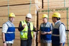 Four people stand in front of tall pallets of wood. Each of them is earing a safety vest and hard hat.