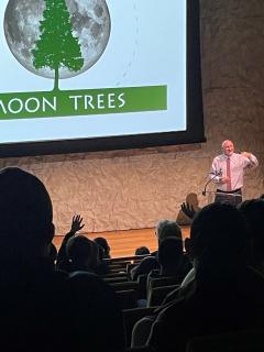 In front of darkened auditorium, Forest Service scientist stands behind a clear lectern. Behind him is a screen with an image of a tree in front of the moon and text "Moon Trees" below.