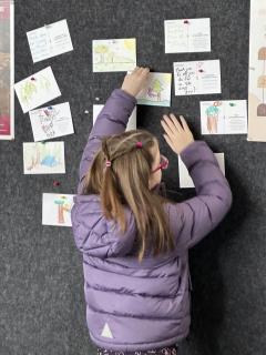 From behind: A child in a purple puffer coat and glasses pins a postcard to a bulletin board.