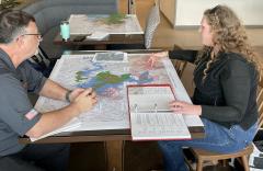 A man and a woman sit on either side of a table with a map spread out between them. The woman is pointing to areas on the map as the man looks on. 