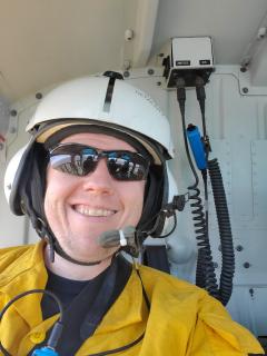 Scott Brownlow in aircraft, wearing helmet, sunglasses, and fire safety clothing. He also has headphones and a microphone.