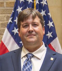 Jeff Marsolais, wearing white shirt, blue patterned tie, suit jacket and Forest Service insignia lapel pin, in front of American flag.