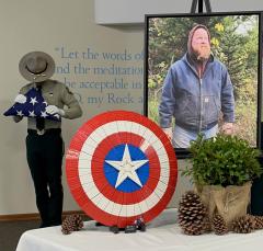 Photo of Casey Budlong next to a Lego Captain America shield. In the background, uniformed Forest Service employee holds a folded American flag to be presented to Budlong's family.