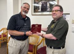 Two men stand in a room with chairs and framed photo of waterfall on wall behind them. Between, them, they hold a small wooden plaque engraved with Forest service insignia and text: Forest Service Safety Award 2023, individual award, Doug Rammer.