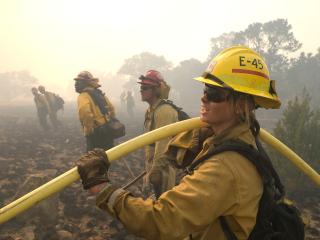 A Forest Service employee wearing safety gear stands in the forefront holding a hose. Other firefighters stand in the background looking at something off camera.