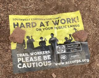 A picture of a flyer on the ground that says, Southwest Conservation Corps, Hard at Work! on your public lands, trail workers, please be cautious.