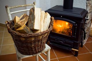 A basket of cut firewood sitting on a wood chair in front of a wood stove. A fire is visible through the glass face of the wood stove door.