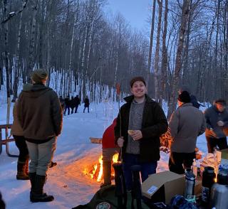 People gather around a bonfire to roast hotdogs and marshmallows. In the background, hikers continue along the snow-covered trail.