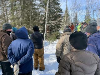 From behind, a group of people in cold-weather gear (coats, snow hats, gloves and mittens) gather in the forest to listen to a speaker.