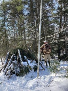 A person in tall winter boots, heavy coat and baseball cap stands in profile in snow. They are next to a pile of cut balsam fir.