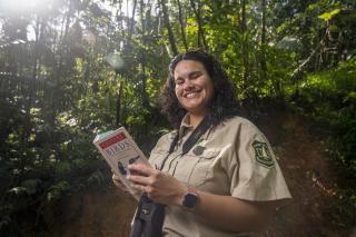 An employee in the forest holding a birding book and wearing binoculars.