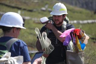 Noah Kaufman carries mesh netting to protect future seedlings and has a GPS antenna on one shoulder and a sack of colored flags on the other. He consults with Jessica Wright before heading out to plant in the burn site.