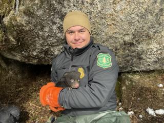 Man wearing a beenie, a jacket with a FS insignia and orange heavy winter gloves holds a black bear cyb in his arms.