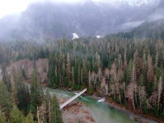 An aerial image of the newly repaired Big Four Ice Caves bridge spanning the South Fork Stillaguamish river. Heavily wooded forest appears on both sides of the bridge and river.