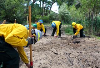 Wildland firefighter trainees crouch around a recently extinguished campfire to search for heat with bare hands and long-handled hand tools.