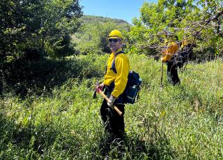 A Forest Service wildland firefighter stands smiling in a forest clearing holding an axe.