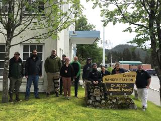 A group of people (11 folks) stand around a sign (that reads: Cordova Ranger Station, Chigach National Forest) and smile for a photo. In the background, there are various buildings, and even further back (to the top right) tree tops and a mountain range.
