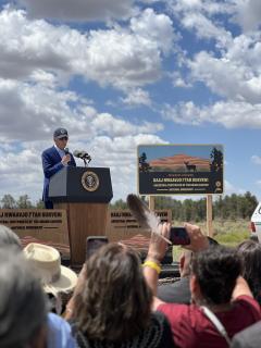 President Biden stands at a podoum, raised on a dais. Next to him (to his left is the sign for the new national monument in the Crand Canyon). At the bottom of the photo, Tribe member are gathered to hear the announcement. 