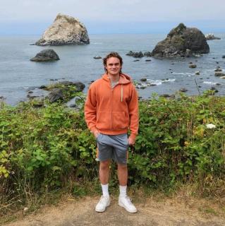 Photo of a young man wearing a faded orange hoodie and grey/blue shorts standing near a coastline. In the far background, where the coast begins, large rocks can be seen jutting out of the water.