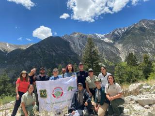 Group photo. Two of the group members are holding a sign with the crests of different groups. The group is standing on a trail, a mountain landscape on the backgroudn.