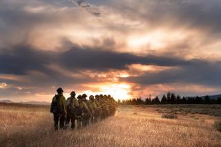 The Wyoming Hotshots walk in a line towards a sunset through a field. 