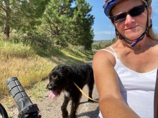 Selfie photo of a woman in a bike helmet, sunglasses, and exercise shirt. Her shaggy, panting dog is on a leash behind her on a trail with pine trees in the background.