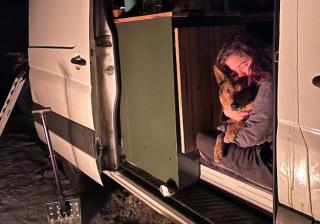 A person holding on to a dog while sitting inside the open side door of a van.