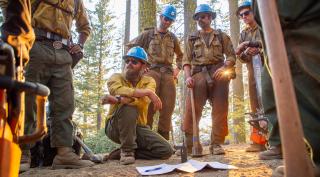 A Hotshot crew standing in a circle surrounding a kneeling firefighter give a briefing in the woods.