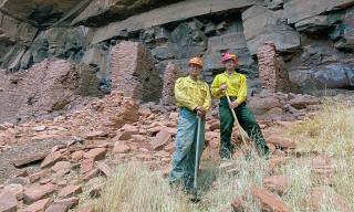 Two wildland firefighters, holding shovels, standing in front of an ancient native american rock structure under a stone cliff.