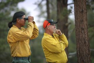 Two wildland firefighters in a forest, observing something off in the distance. One person is using binoculars.