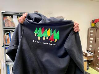 Forest pathologist Martin MacKenzie holding up a hoodie sweatshirt with illustrations of colored trees on the back. At the bottom is the statement "I see dead trees..."