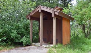 A brown, wooden, connected vault toilet facility that is only big enough for a person to stand in without assistive devices.