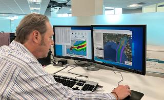 A person at a computer with two screens that show hurricane paths over Florida, one is the national hurricane center model predicting wind speed risk and one is the TreeS-DIP model predicting damage