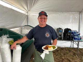 A man holding a disposable plate full of food, smiles for the camera while he reaches for a styrofoam countainer with his other hand