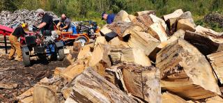 People working next to piles of wood with power log splitters to split cut logs into firewood size pieces.