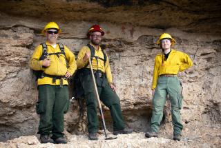 Three uniformed forest service employees stand with contented looks on their faces on a rocky outcropping in front of a wall with painted pictographs.