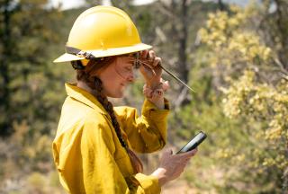 A uniformed forest service employee tips up her sunglasses under her safety helmet as she looks down and enters information on a small electronic device.