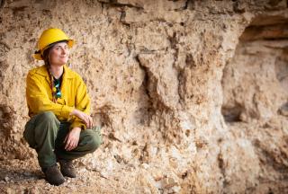 A uniformed forest service employee crouches, smiling, on the ledge on a rocky wall.