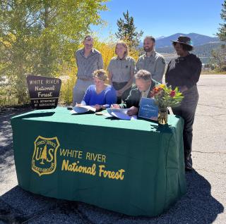 Two people signing agreement at a table with table cloth that says White River National Forest. 