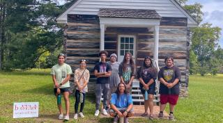 Students stand in front of renovated log cabin with historical significance.