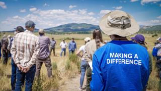 A man wearing a blue long sleeved t-shits with the words “Making a Difference” on the black, stand in an open field with several other people listening to a speaker talk with green mountains in the background and puffy clouds in the sky.