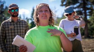 A woman wearing a bright neon green t-shirt, holding a paper with her hand on her chest gesturing with aa smile on her face. Two individuals over each shoulder, standing in the background, listening to her share information.