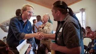 Two people from different cultures smiling and shaking hands in a crowded recreation room.