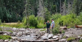 A Native American Man wearing a green polo shirt is standing on the bank of a rocky creek with rustling water next to a white man with a ball cap and short-sleeved shirt, pointing out over green shrubs and trees that have come back after a somewhat recent fire.