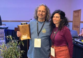 Two people stand together smiling, in a conference, with potted river cane beside them.