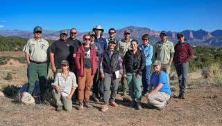 A group of people pose in front of green woodlands and red rock mountains behind them.