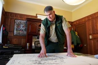 Employee in uniform in a wood-paneled office. He stands over a map of oil and gas wells spread across a table.