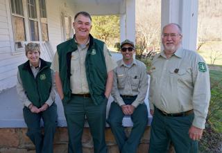 Group photo, employees in uniforms on edge of porch to ranger district office. Left to right: Trish Boles, recreation manager; Bobby Claybrook, district ranger; Wes Sprinkle, silviculturist; and David Breetzke, archeologist.