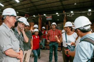People wearing hard hats standing in a loose circle in a warehouse with stacks of wood in the background.